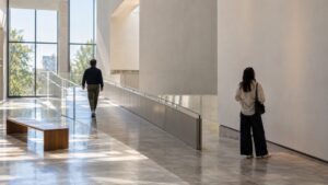 Two visitors moving and pausing in a bright museum lobby beside a sloping ramp